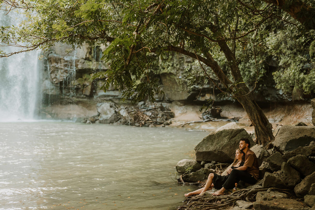 costa rica An engagement photographer captures beautiful engagement photos of a woman sitting on a rock next to a waterfall in Costa Rica.