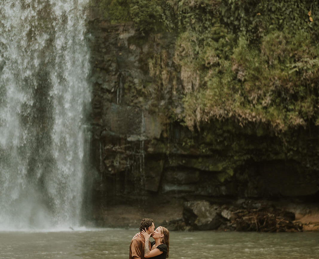 costa rica A couple capturing their engagement photos in front of the stunning Top Waterfall in Costa Rica, sharing a passionate kiss.