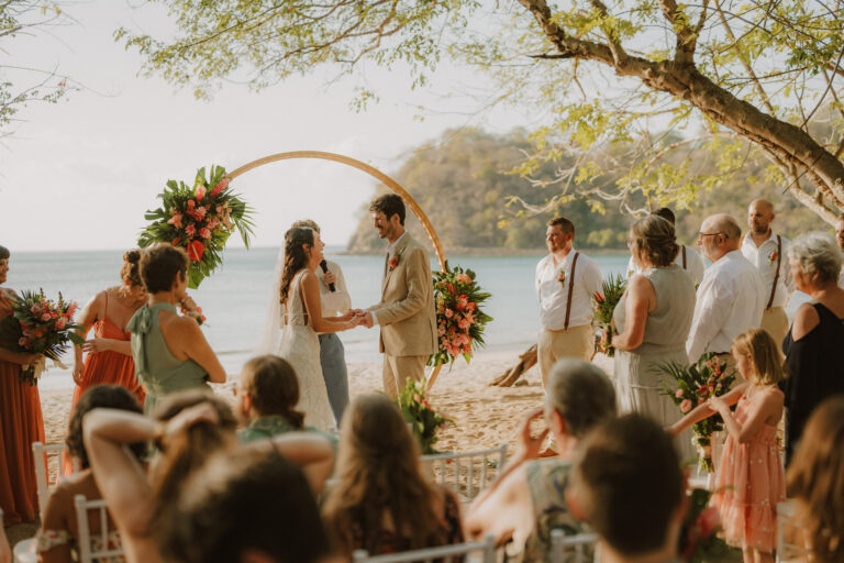 costa rica Dreams take shape as April and Bryan exchange vows under a floral arch on the beach, surrounded by guests in semi-formal attire.