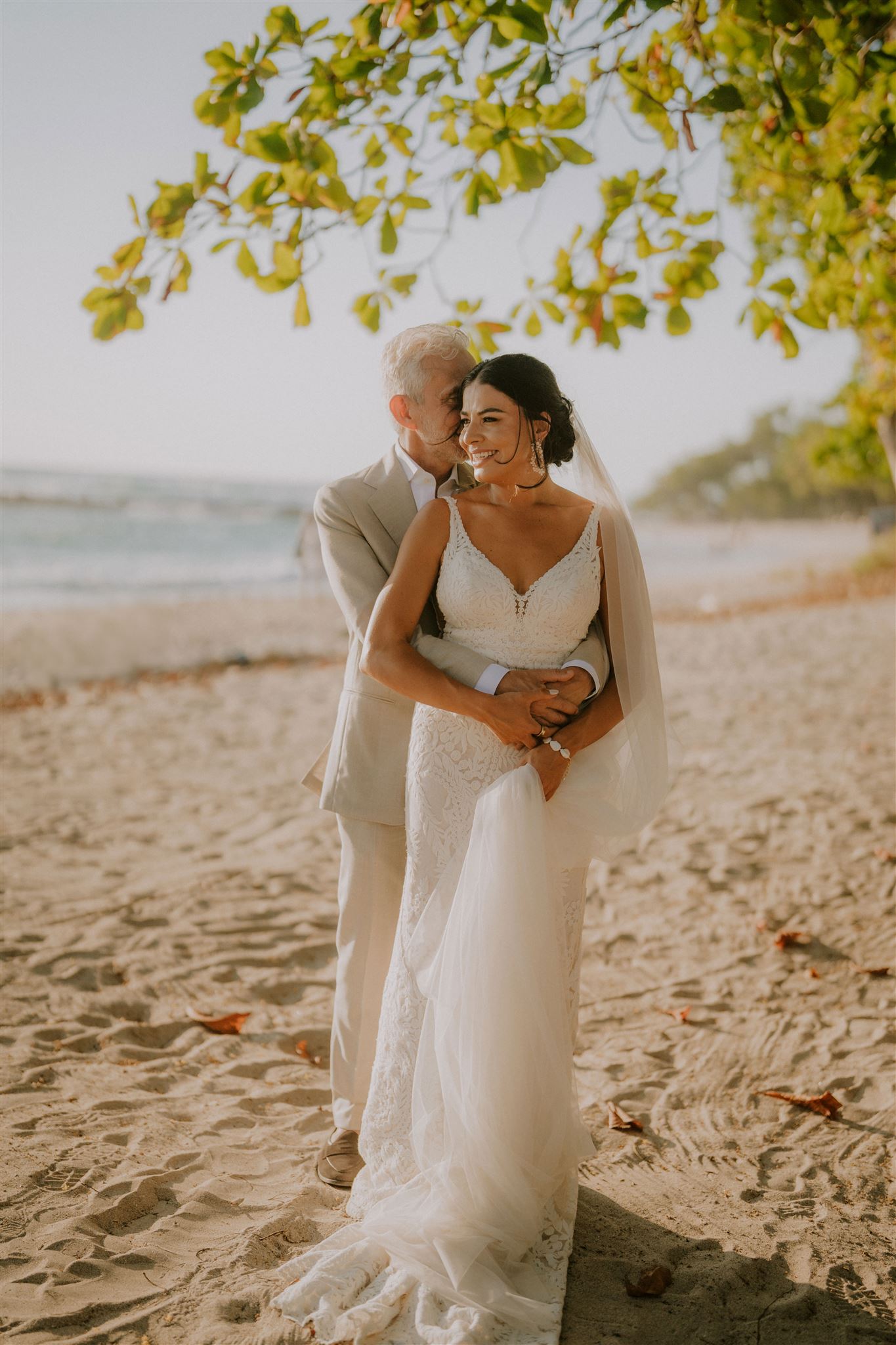 costa rica A bride and groom stand on a sandy beach under a tree, embracing and smiling, with the ocean and greenery in the background. Discover the magic of their love story against nature’s breathtaking backdrop.