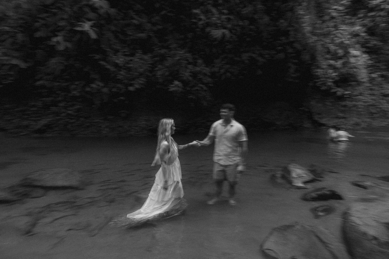 A couple crosses river rocks hand in hand in black and white, rainforest rising behind them at Amor Arenal