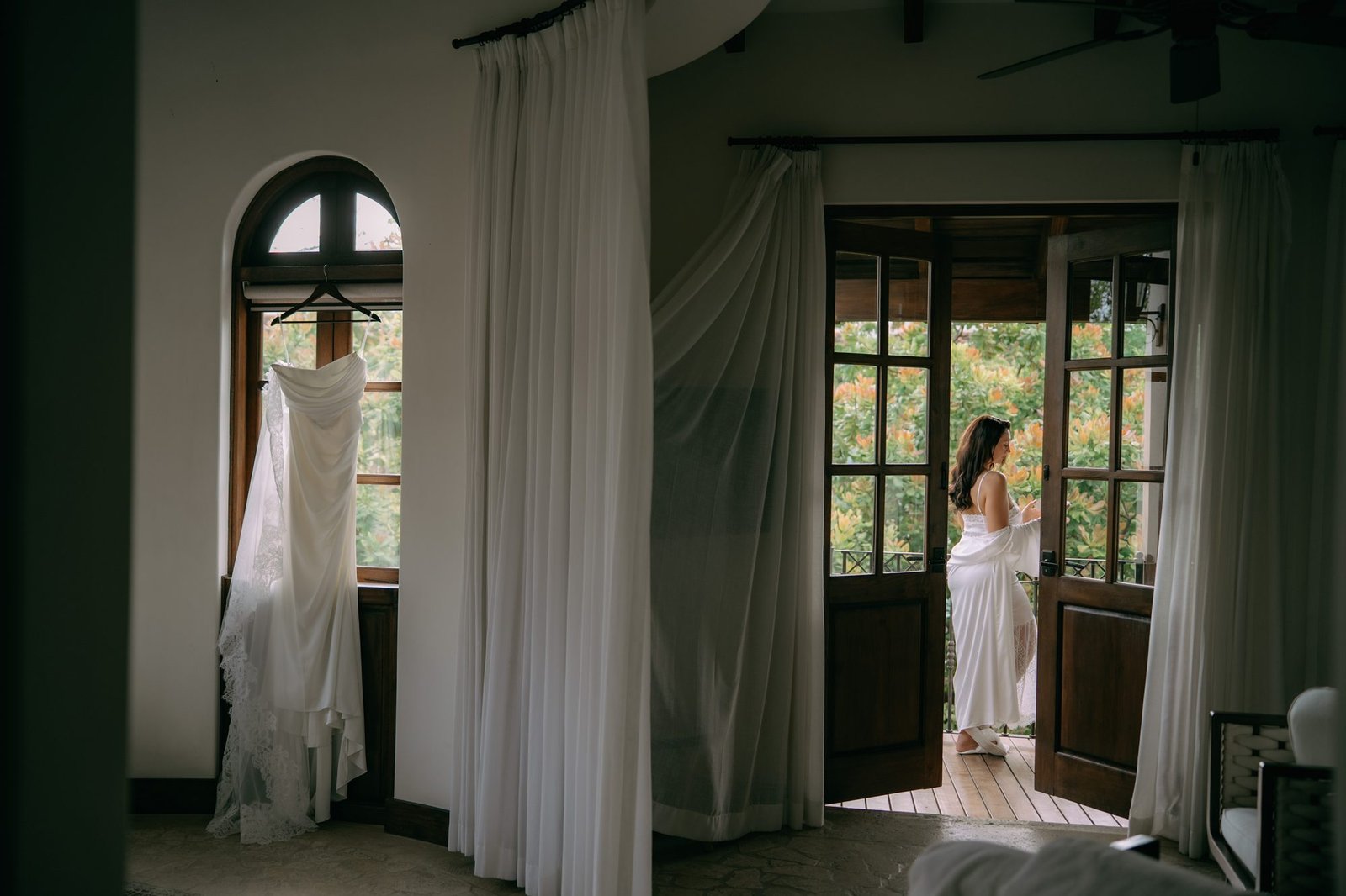 A wedding dress hangs in an open doorway while the bride stands in the room beyond