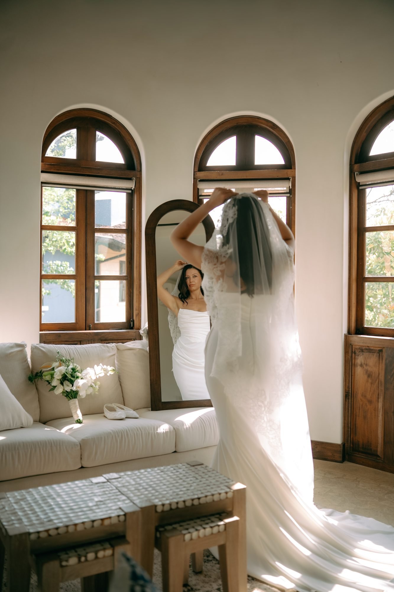 A bride lifts her veil beside a tall window, morning light falling across her dress