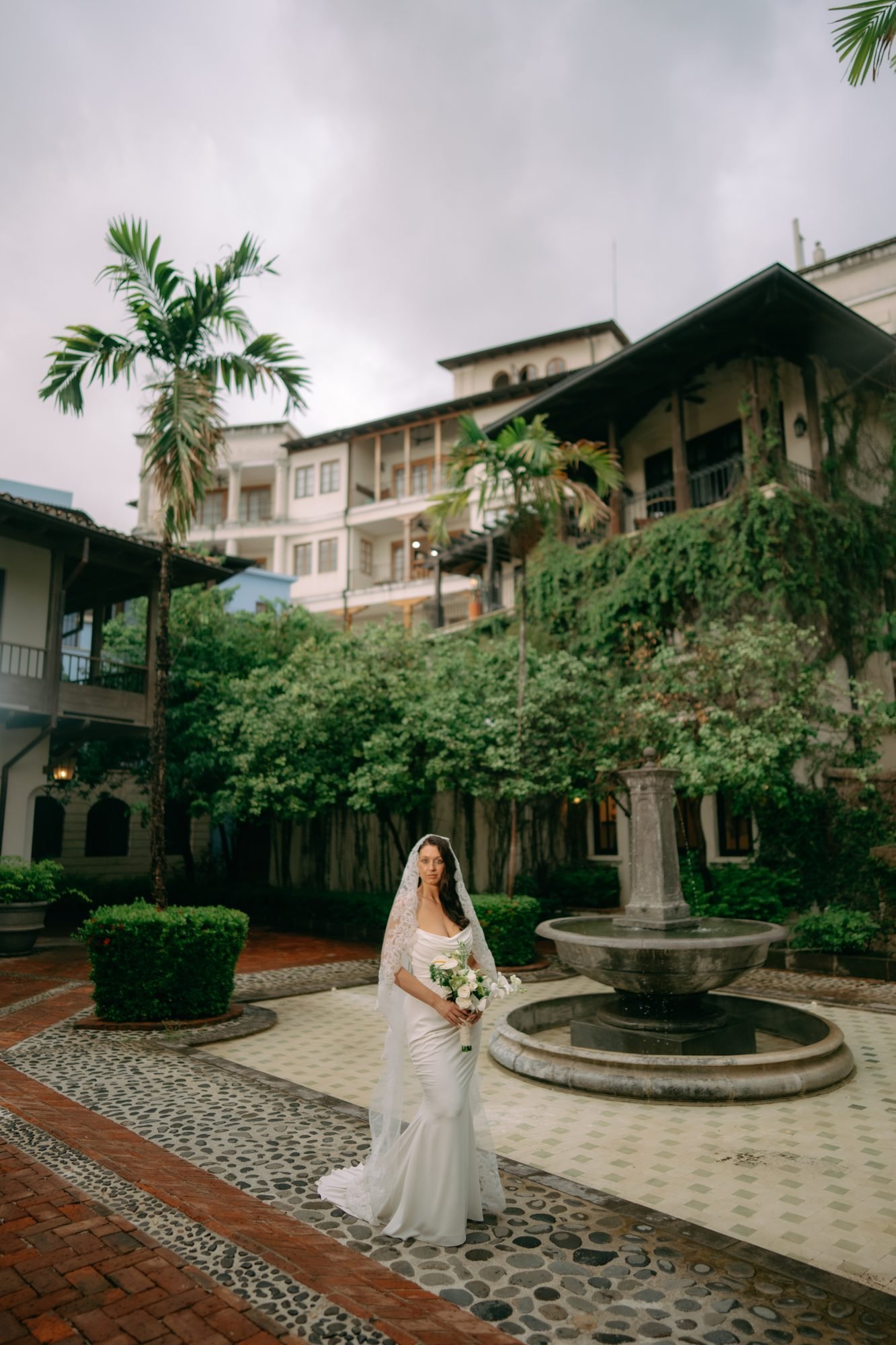 A bride walks across a colonial courtyard toward a stone fountain, arched buildings and palms around her