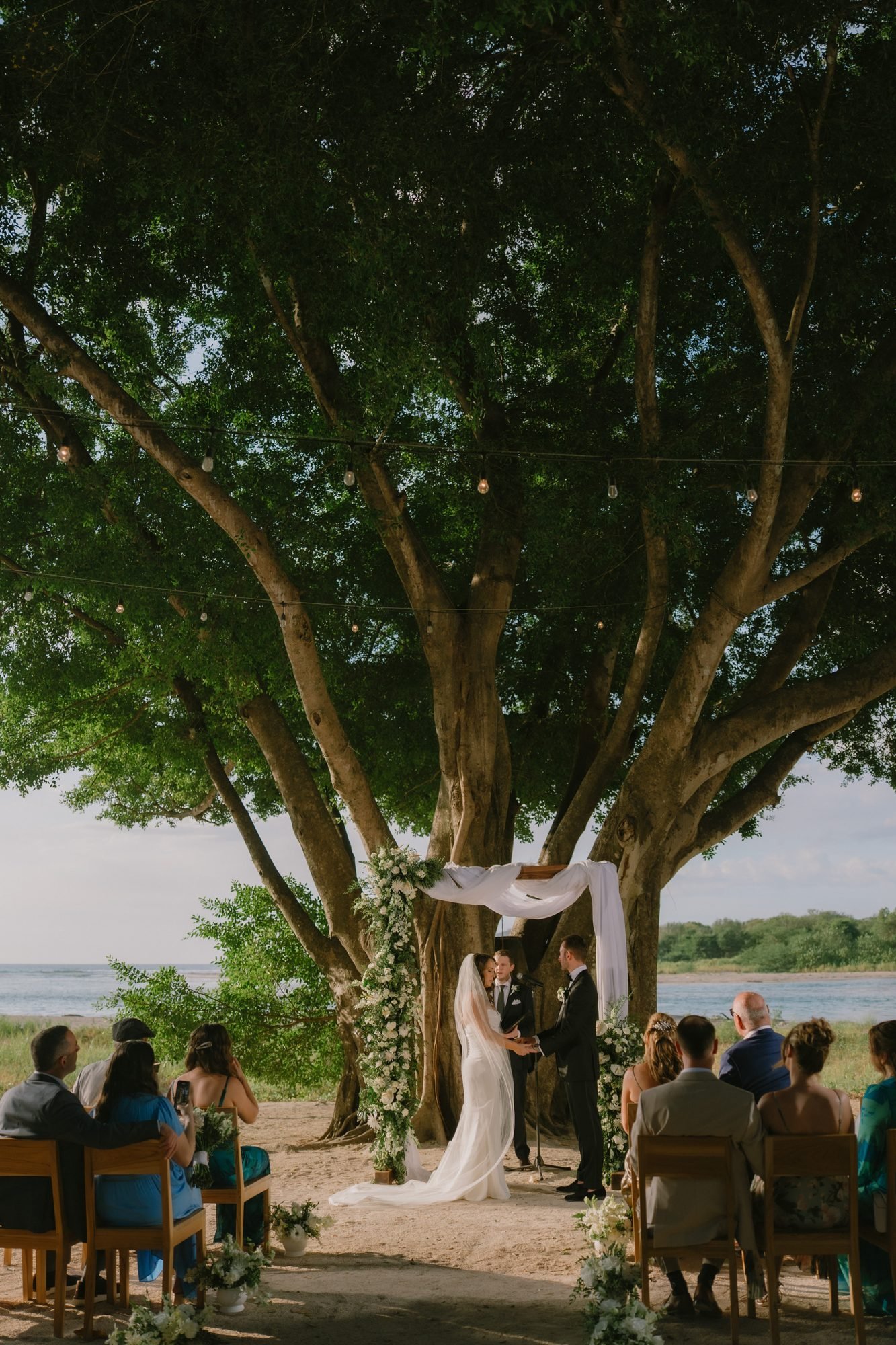 A small wedding ceremony takes place under a large spreading tree beside water, with a handful of guests seated