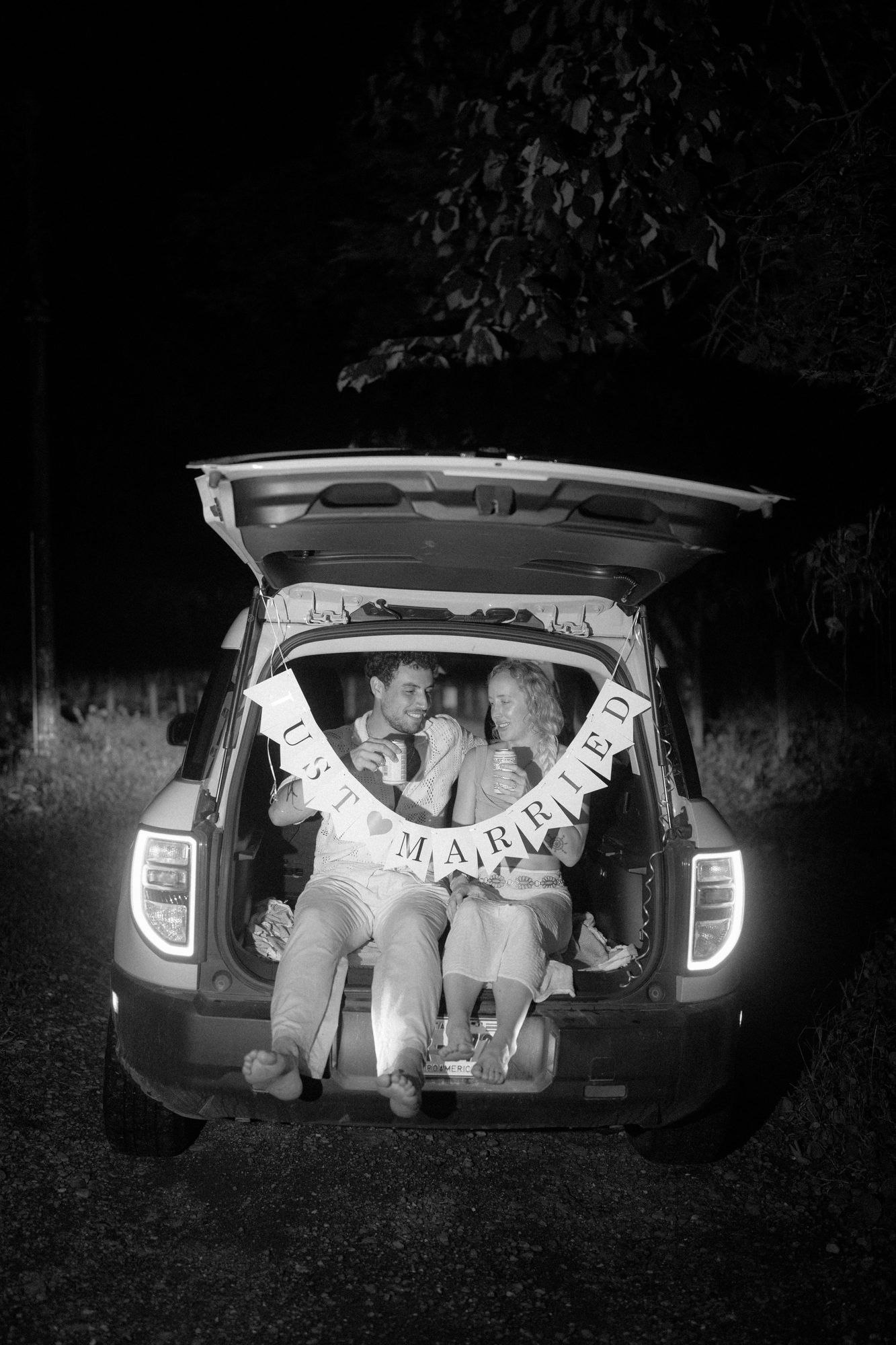 In black and white at night, a group sits in the open boot of a car under strings of lights, photographed by a Costa Rica elopement photographer