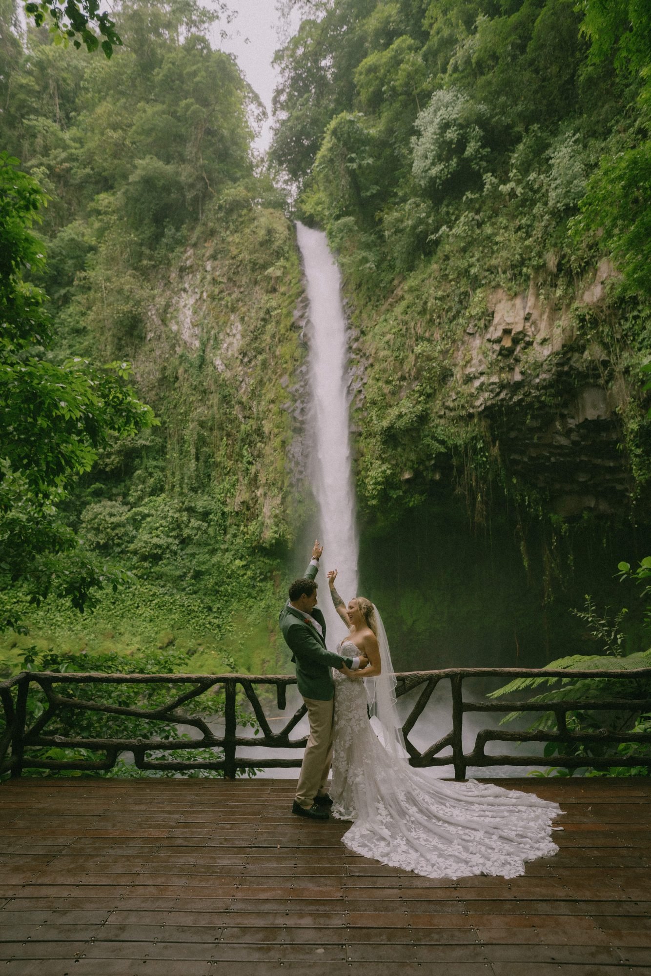 A couple stands together on a wooden platform facing a tall jungle waterfall in Costa Rica