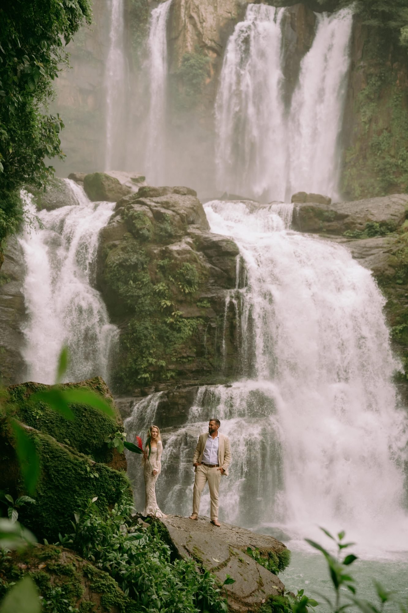 A couple stands small at the base of a wide multi-tiered waterfall surrounded by rainforest