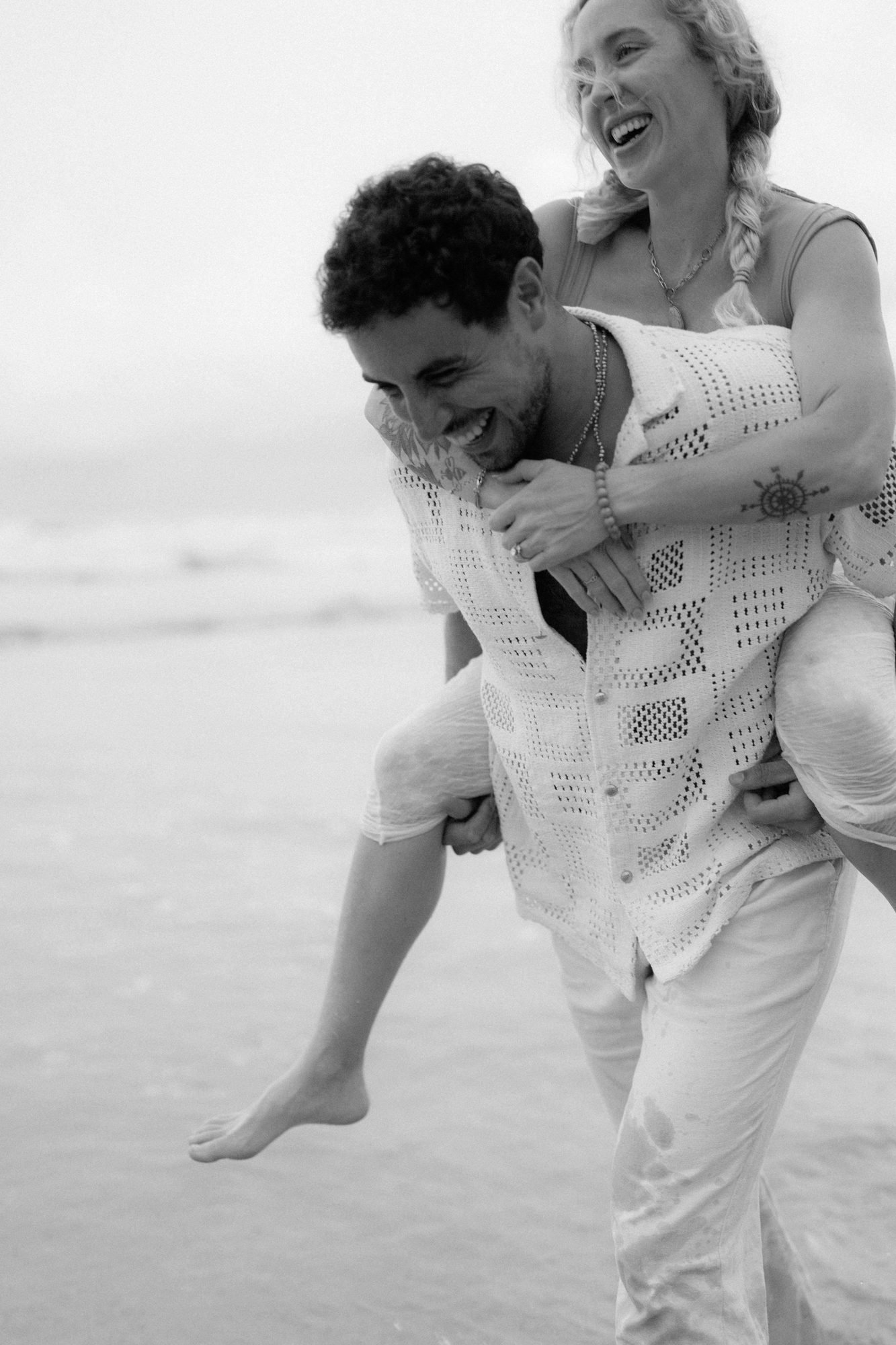 In black and white, one partner carries the other along the beach as they both laugh