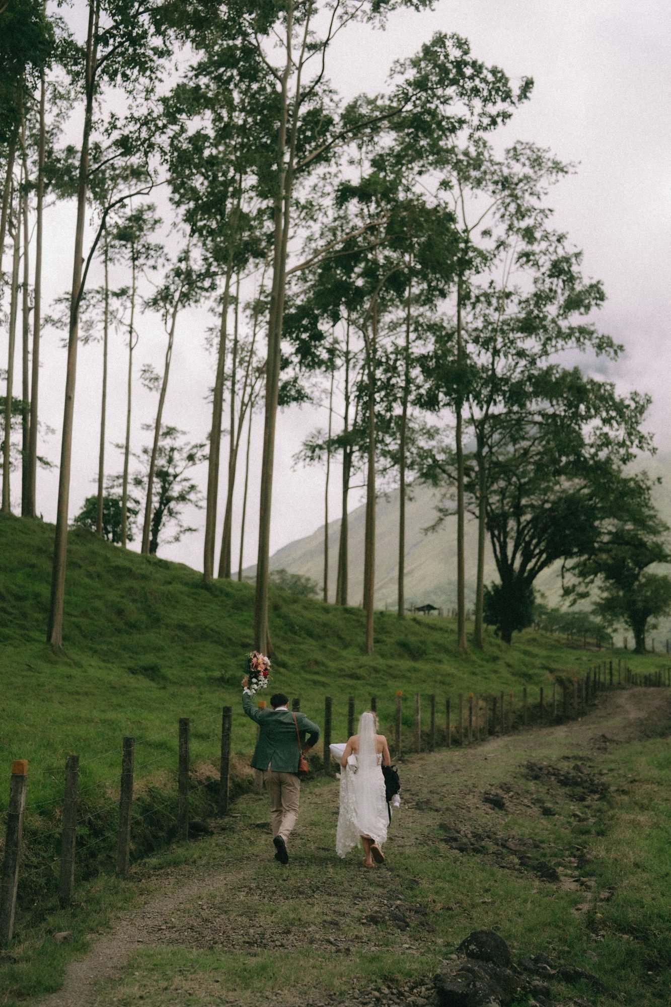 A couple walks a narrow path along a green ridge with mist rising through the trees behind them, photographed by a Costa Rica elopement photographer