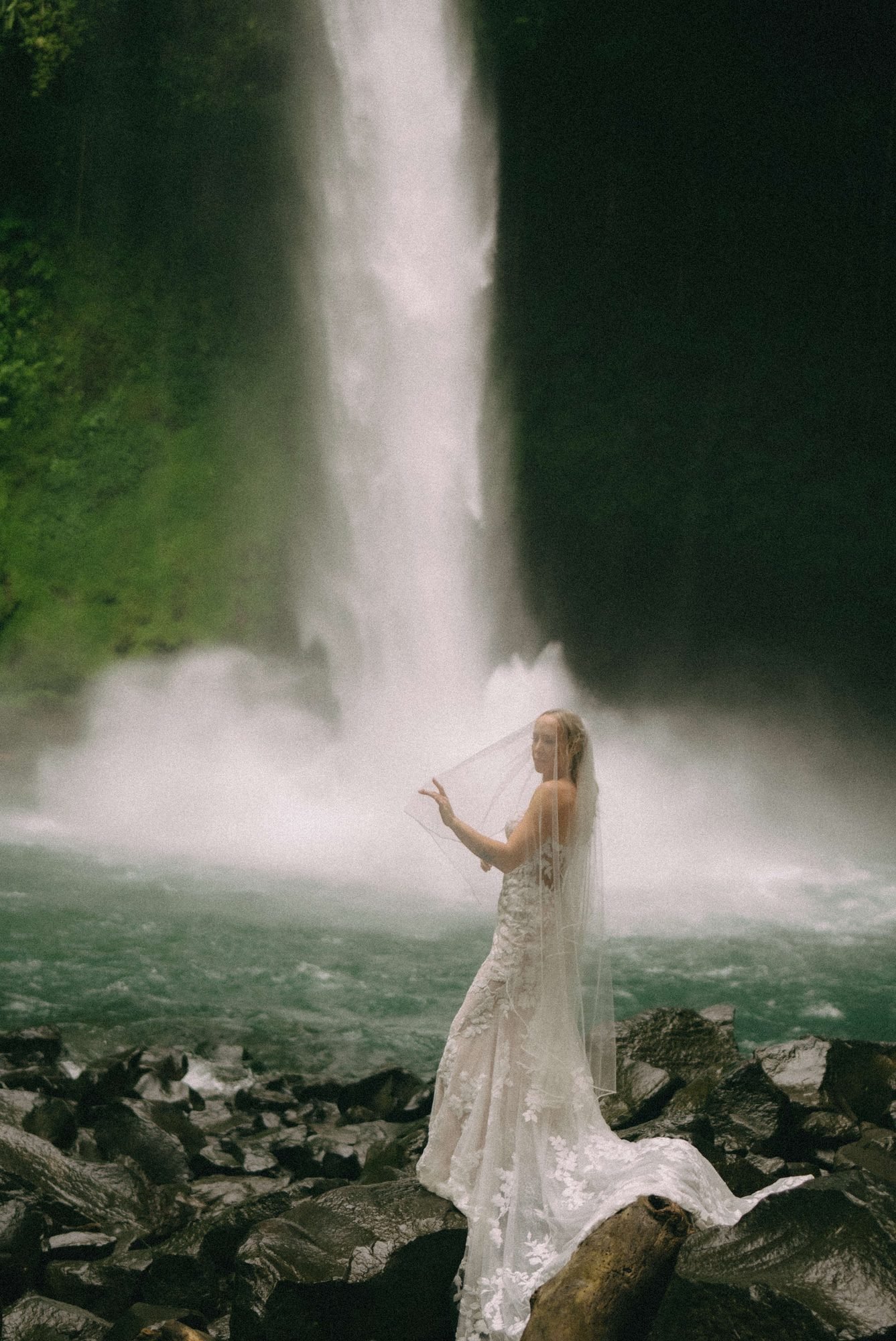 A bride stands alone on rocks at the base of a tall waterfall, spray rising around her