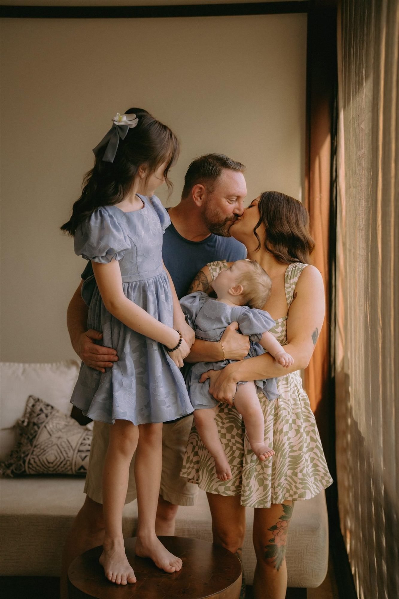 A family with two young children stands together in soft window light before a wedding