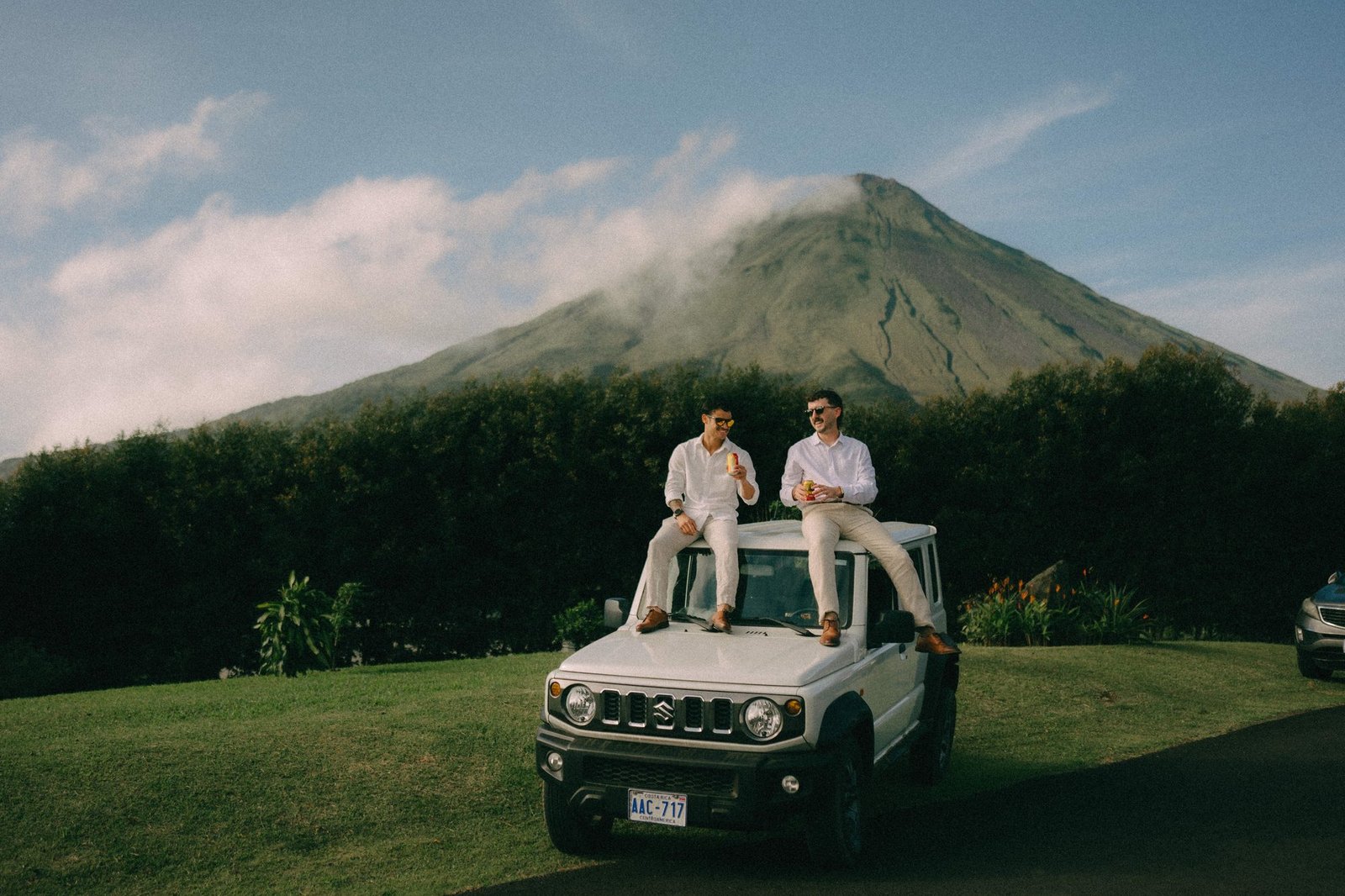 A couple sits on the bonnet of a four wheel drive parked on grass with Arenal Volcano behind them