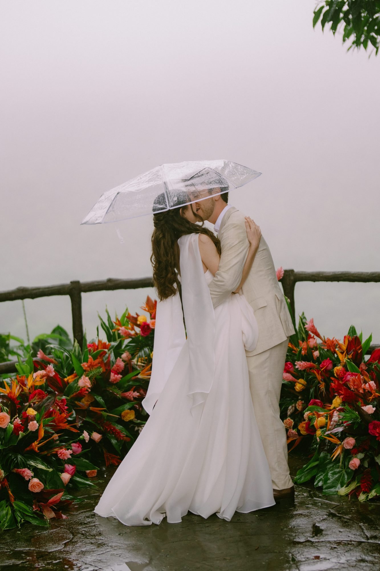 A couple stands close together under a clear umbrella after their ceremony, rain falling on tropical flowers behind them