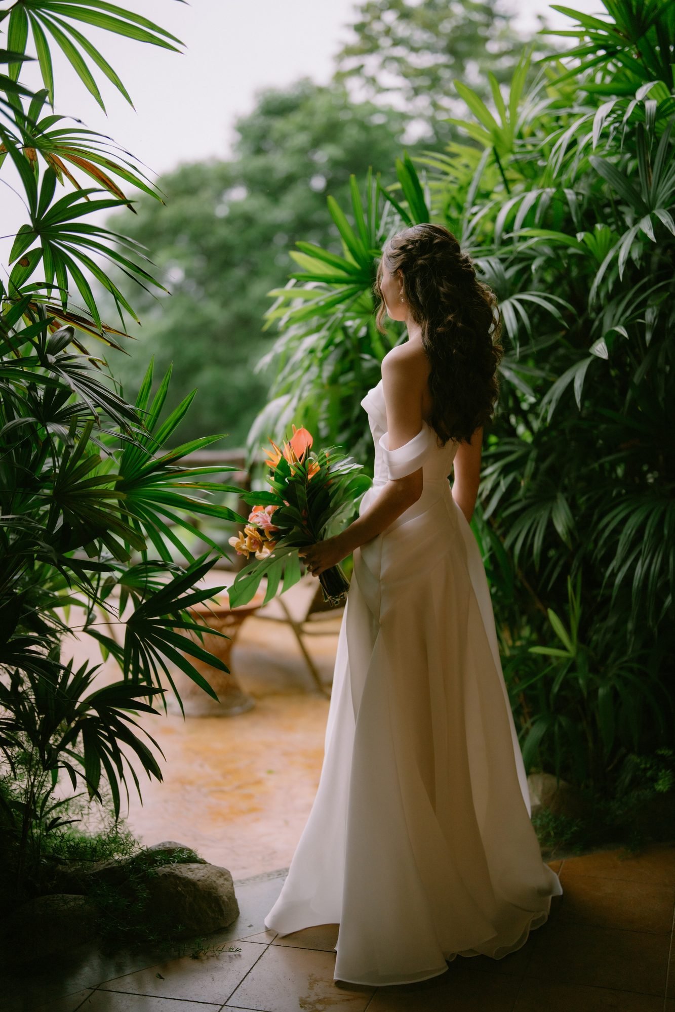 A bride stands among tropical foliage holding a bouquet, warm light falling through the leaves
