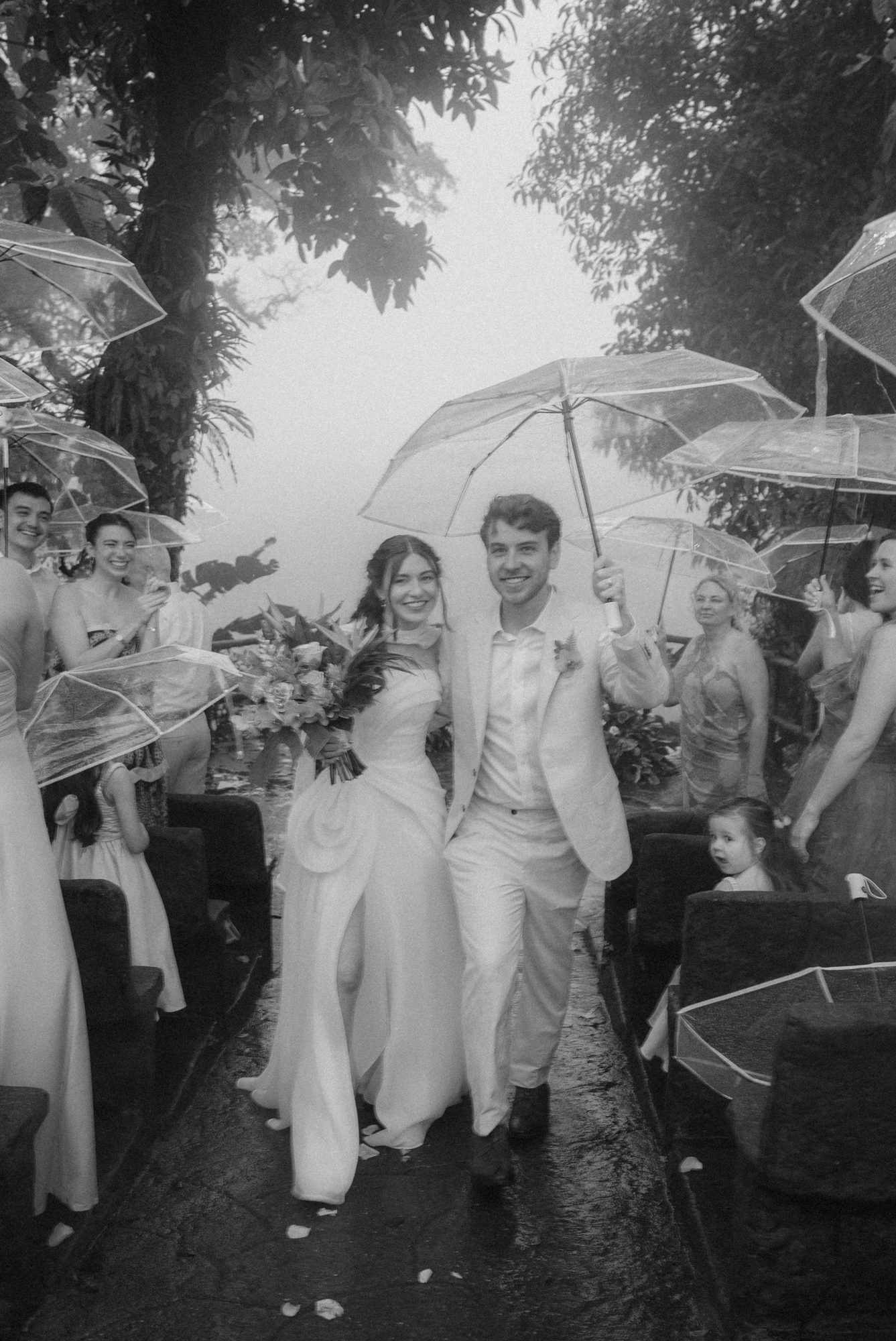 Guests cheer under umbrellas as a newly married couple walks back down the aisle in the rain