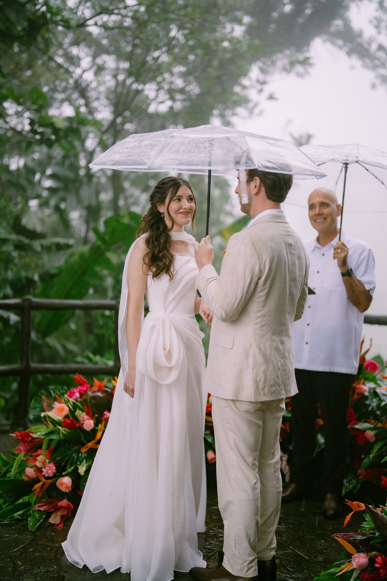 A couple stands with their officiant during a rainy ceremony beside an arrangement of red and orange flowers