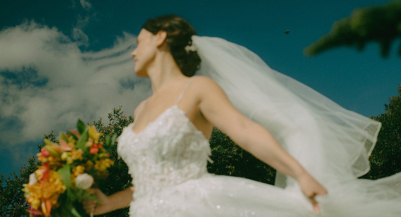 A bride turns with her veil lifting behind her, holding a tropical bouquet against a blue sky