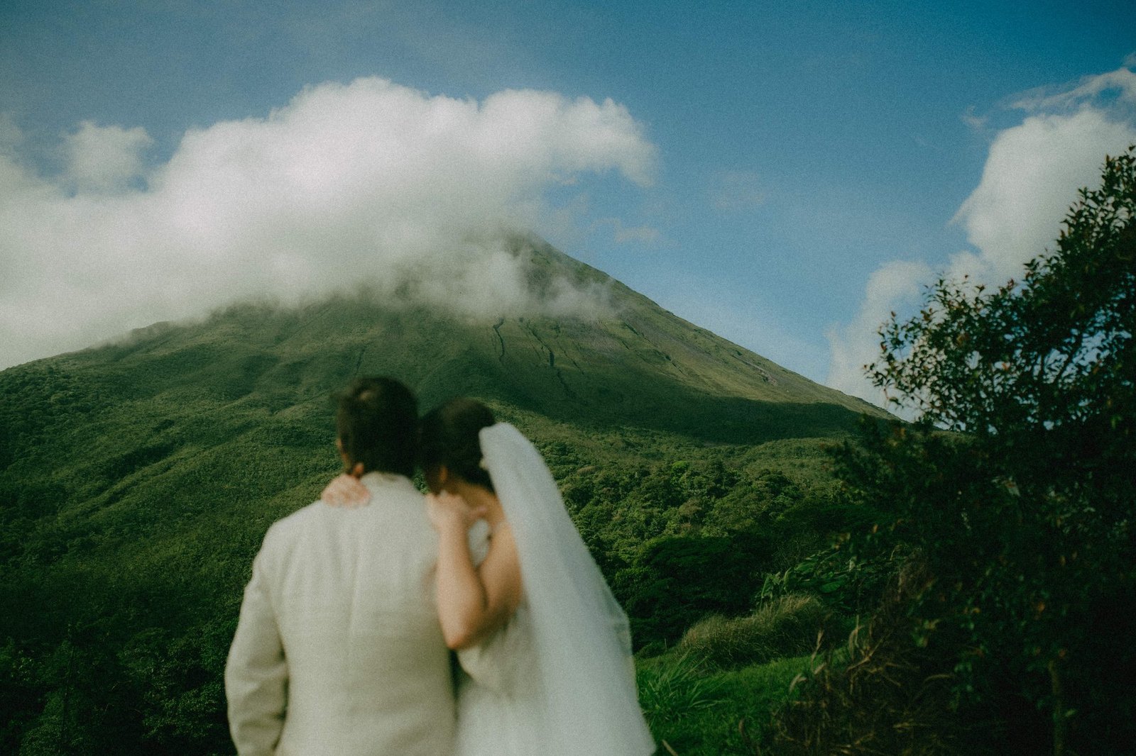 A couple stands with their backs to the camera looking up at Arenal Volcano, its peak wrapped in cloud, the bride's veil falling over her shoulder, photographed by a Costa Rica elopement photographer