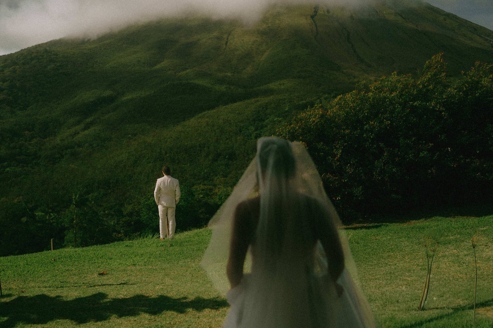 A bride's veil blows across the foreground as her partner waits in the distance on a green hillside
