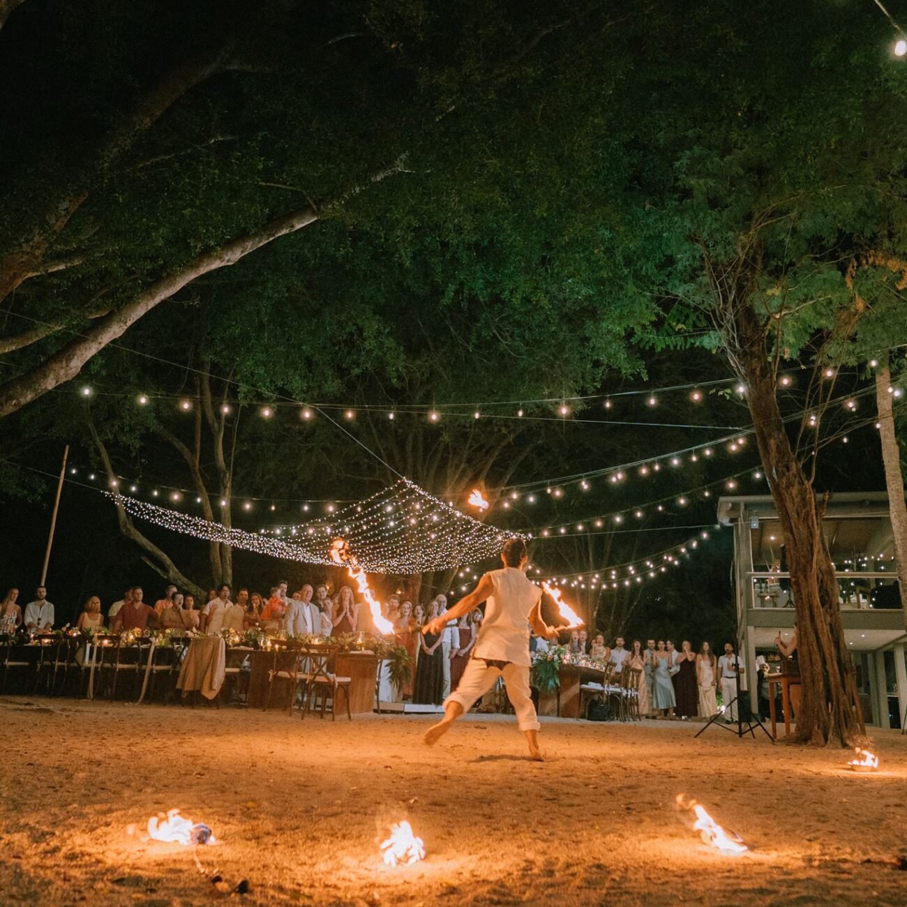 A fire dancer performing on the sand under the trees at Pangas Beach Club, guests seated at long tables behind