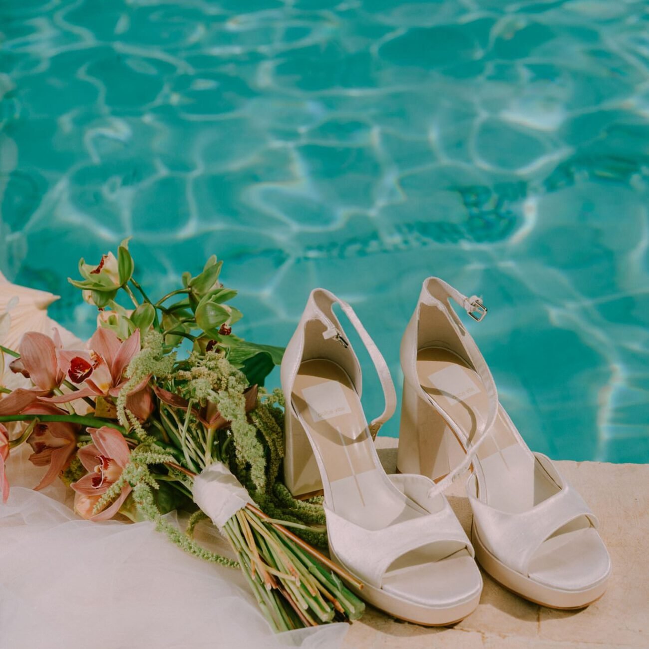Bridal shoes and a pink and green bouquet at the edge of a pool
