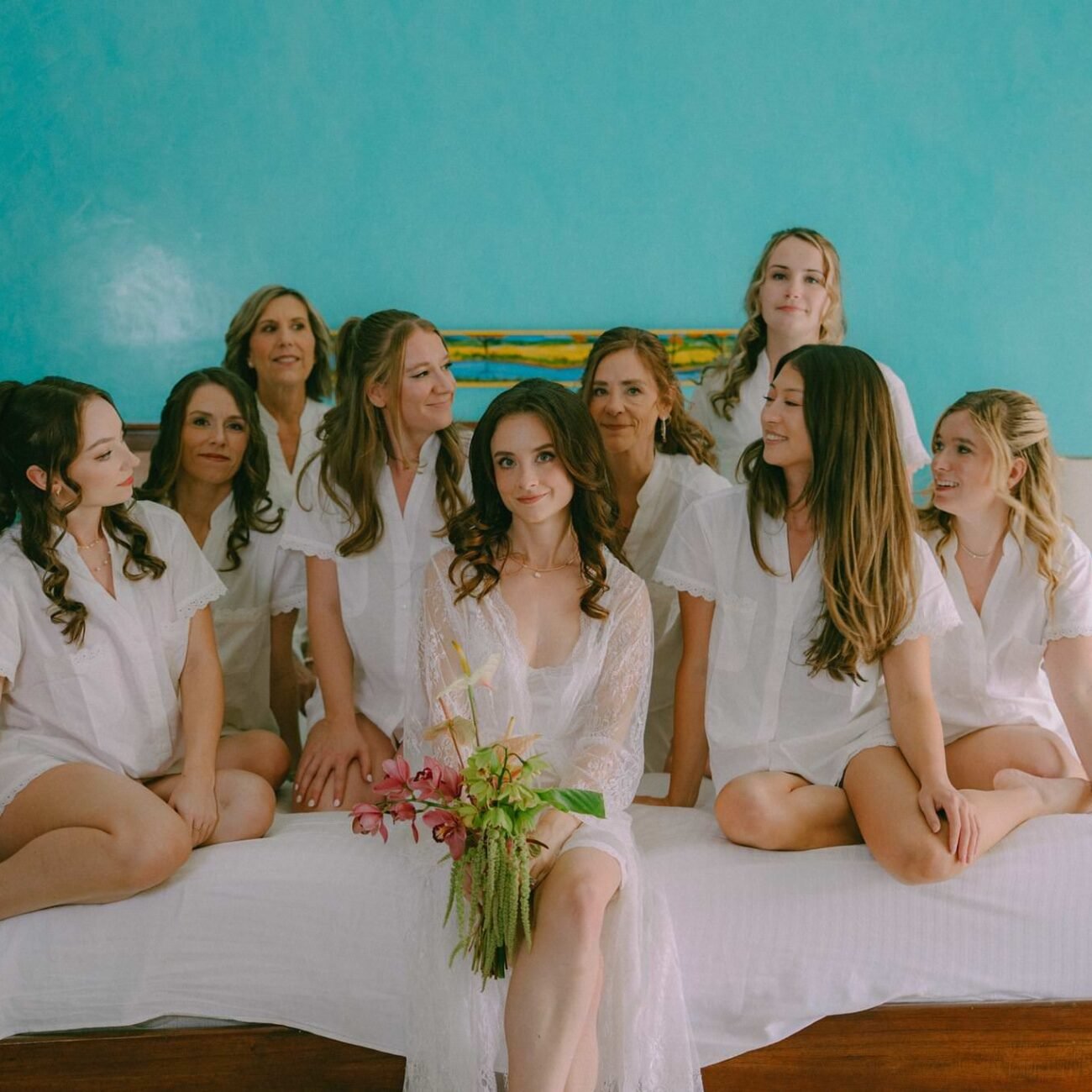 The bride and her best friends on a bed in white pajamas against a turquoise wall