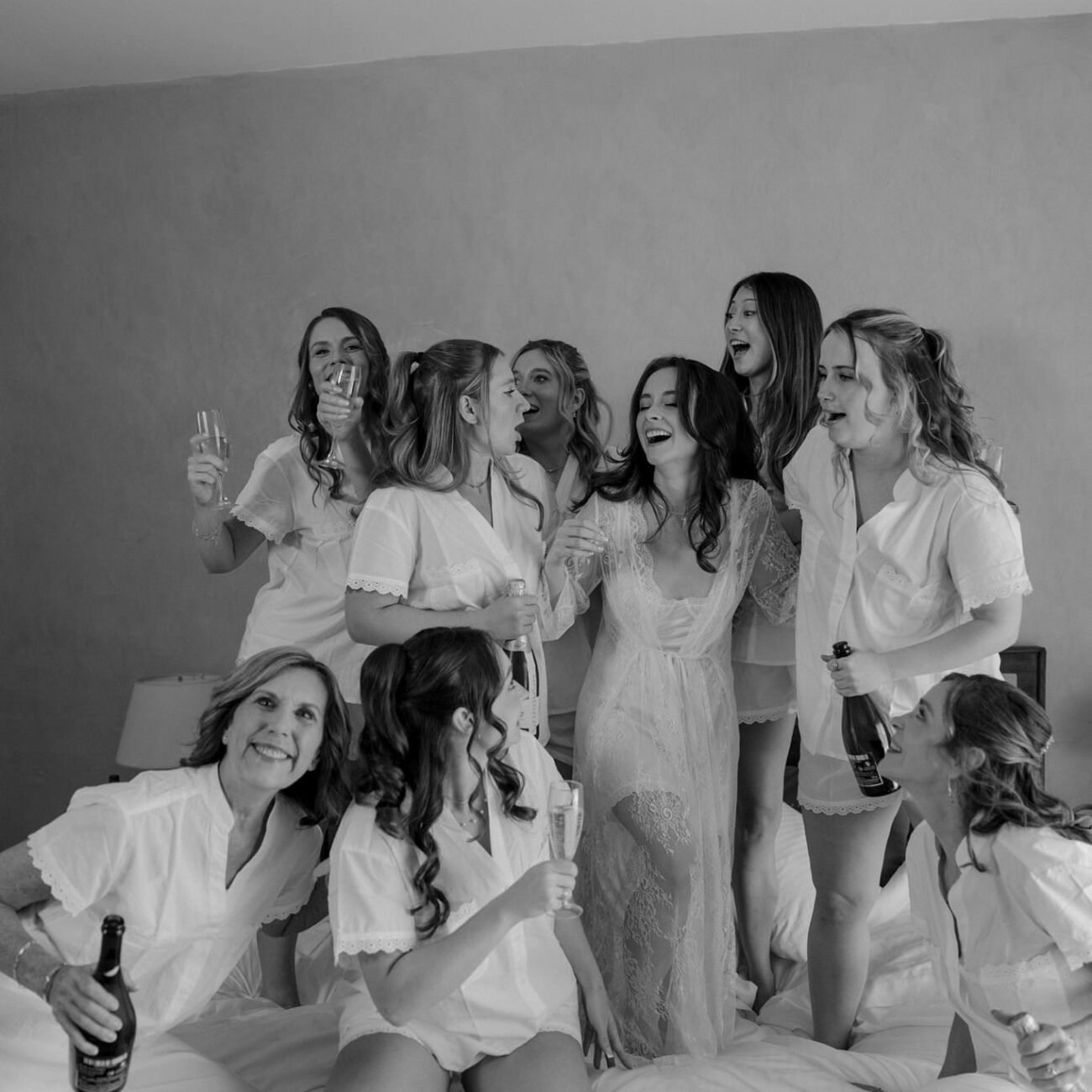 Friends in white pajamas laughing and jumping on the bed while the bride gets ready