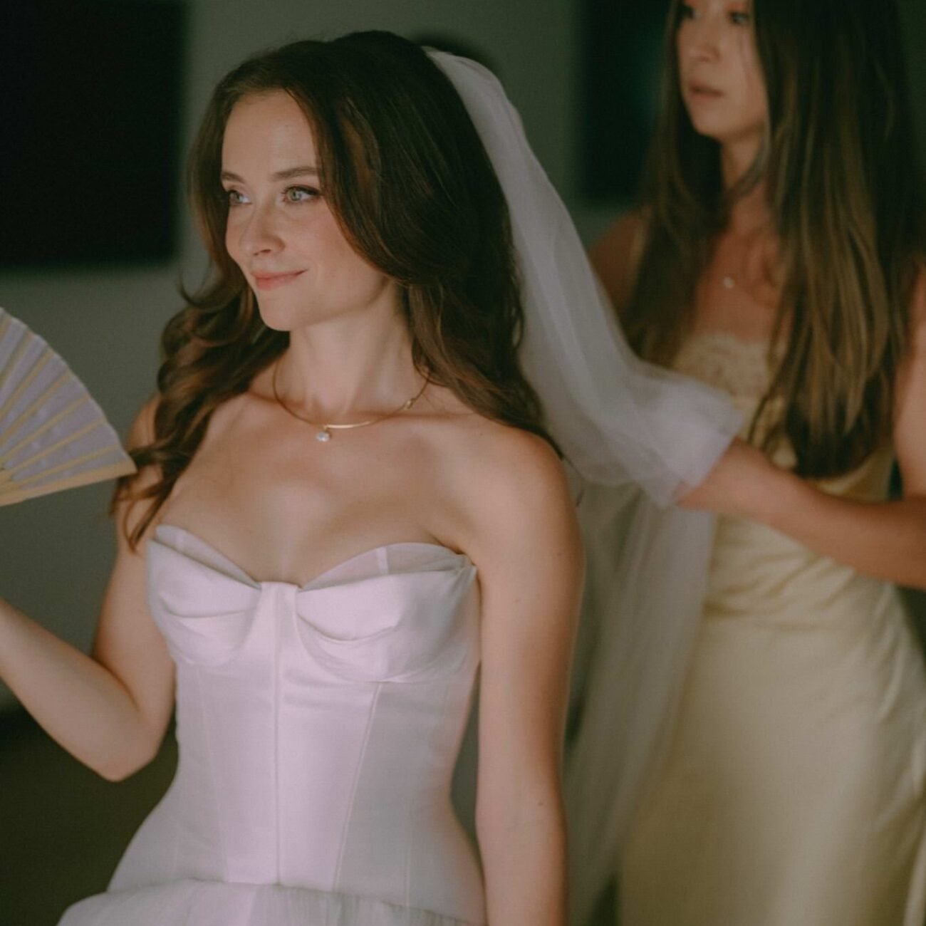 The bride in her wedding dress while a friend arranges her veil, holding a hand fan