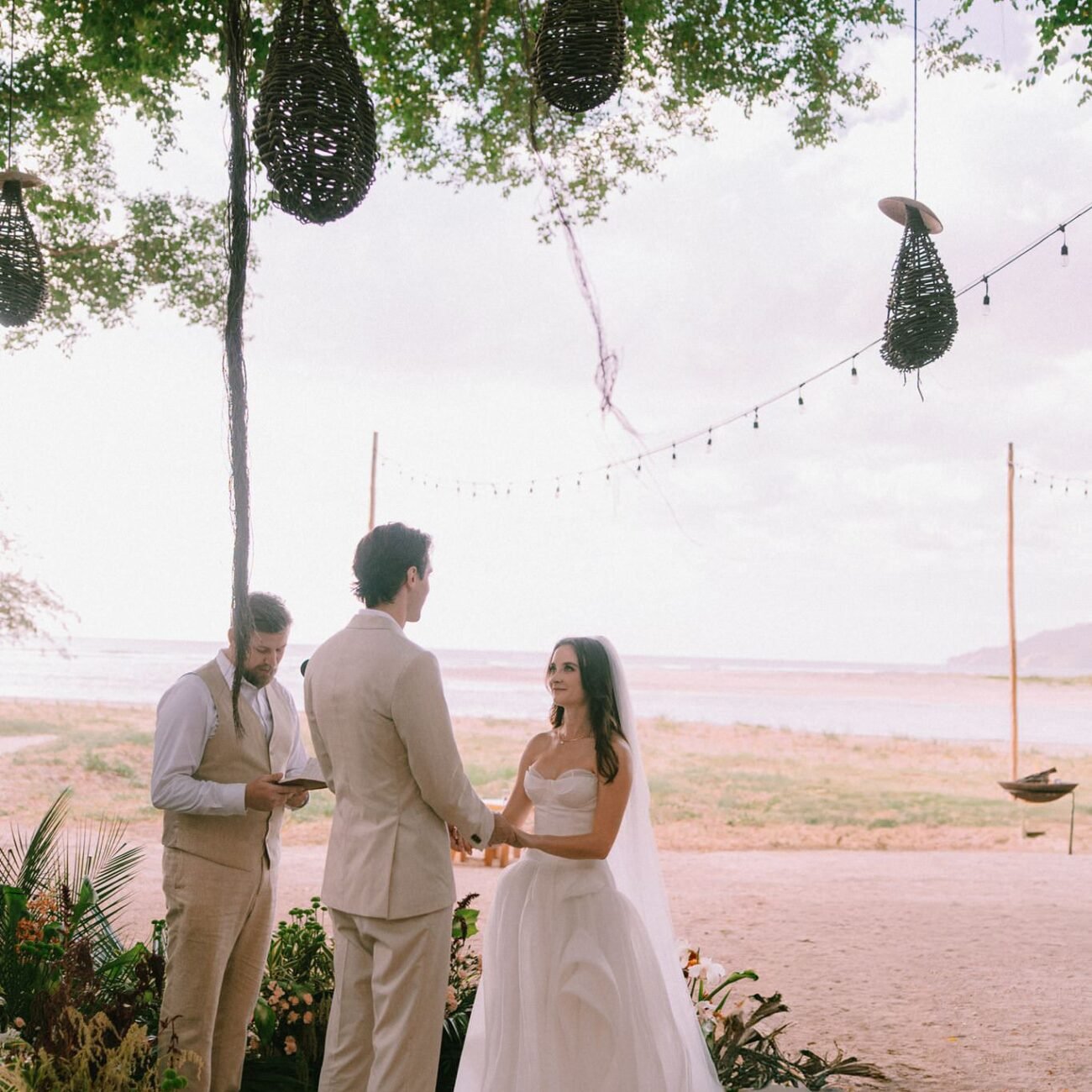 The couple with their officiant at the ceremony, a tree with hanging lights above them and the ocean behind