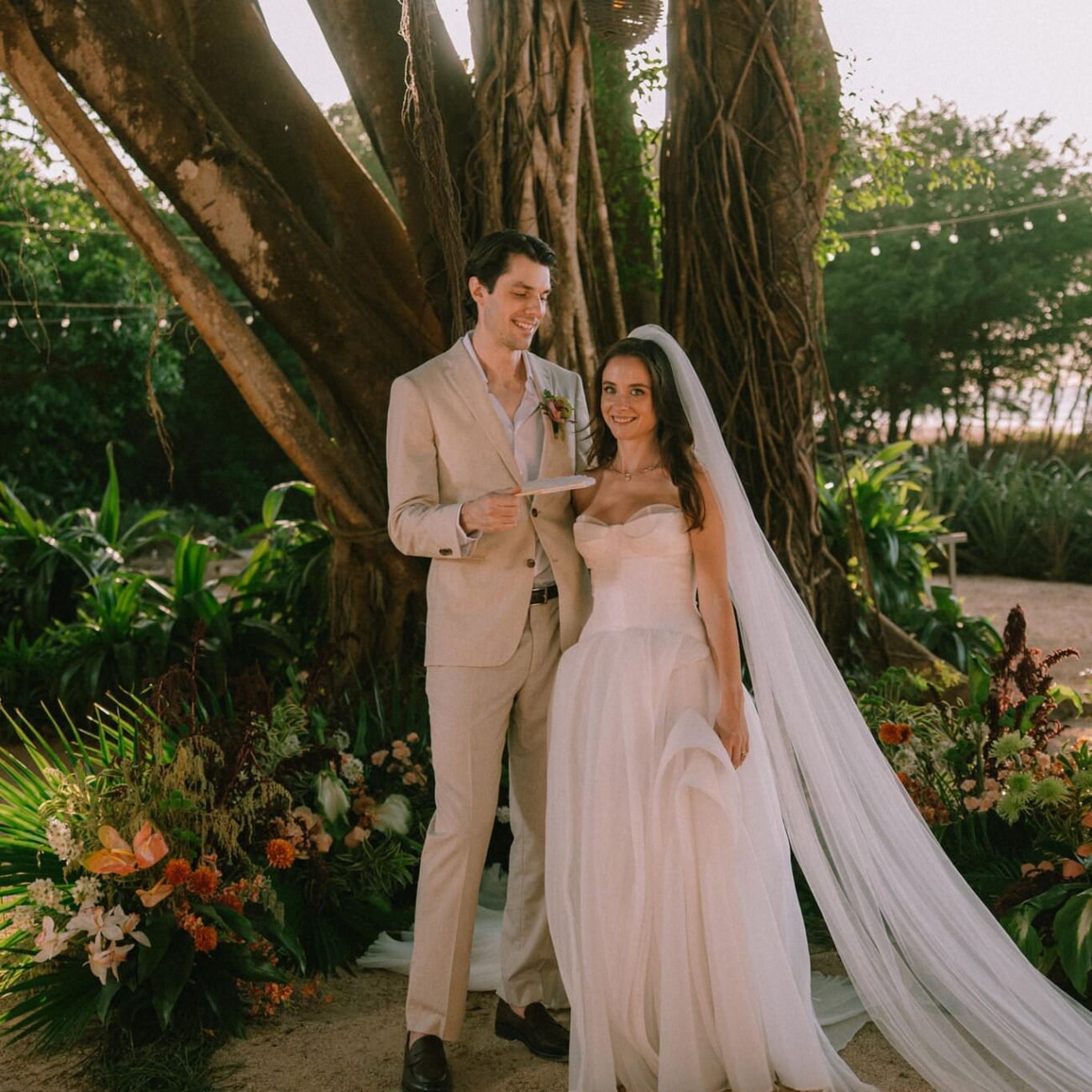 The couple standing together under a large tree, the bride’s long veil trailing