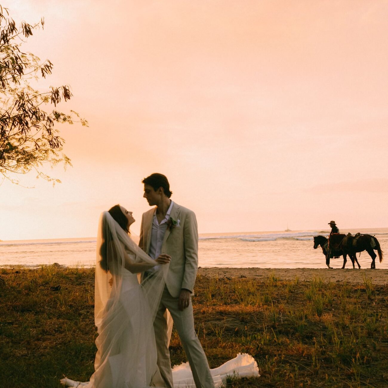The couple embracing on the beach at sunset with two horse riders in the distance
